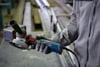 Person in workwear holds a blue and black Bosch angle grinder with a red button in an industrial setting.