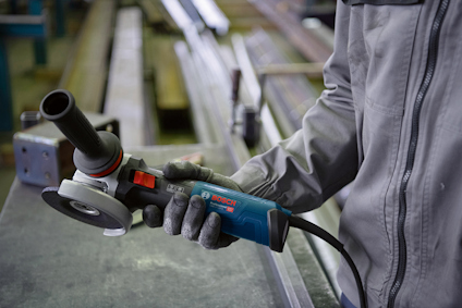 Person in workwear holds a blue and black Bosch angle grinder with a red button in an industrial setting.