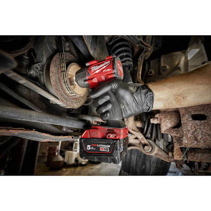 Hand med handske använder ett Milwaukee M18-verktyg på en bils underrede. Rustiga delar syns.