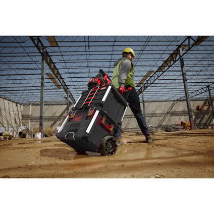 Person in work gear pulling a wheeled toolbox on a construction site. Steel framework visible.