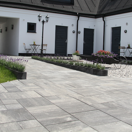 Stensatt innergård med blommor och buskar, tre dörrar och två höga lyktor framför en vit byggnad.