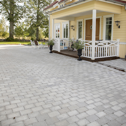 Gult trähus med veranda, omgiven av stenlagd uteplats och träd i bakgrunden.