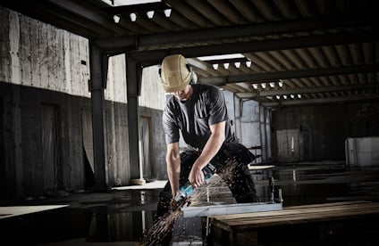 Person i hjälm och skyddsglasögon kapar metall med en vinkelslip i mörk industrilokal.
