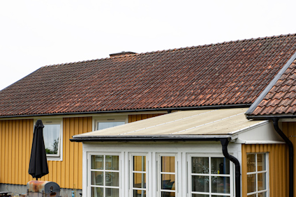 Gul husfasad med svart tak, vita fönster och en glasveranda. Parasoll och stuprör syns.
