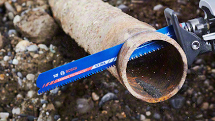 En rostig, cylindrisk metallrör kapas mitt i med ett blått sågblad märkt "Bosch" mot en grusig bakgrund.