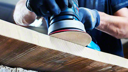 Person slipar träyta med en elektrisk slipmaskin och bär handskar.