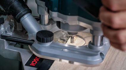 En person använder en fräs för att bearbeta en träyta, med fokus på fräsens spets.