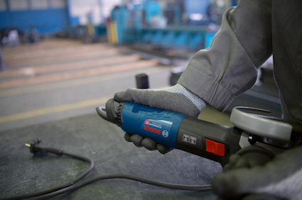 En person håller i en blå bandslipmaskin med röd knapp, klädd i grått med skyddshandskar i en verkstadsmiljö.