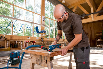 Person slipar träbräda med elverktyg i verkstad, omgiven av verktyg och fönster med gröna träd utanför.