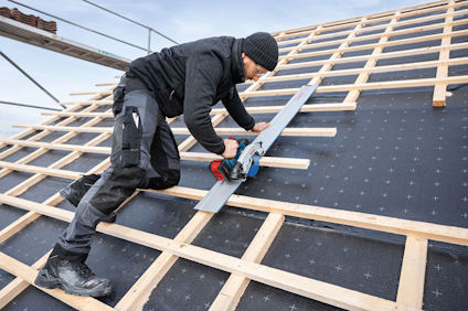 Arbetare i mörka kläder sågar träreglar på ett tak med hjälp av en cirkelsåg. Taket är täckt med svart underlag.