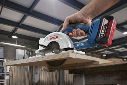 Hand håller en Bosch cirkelsåg som skär genom en träplanka i en fabrik.