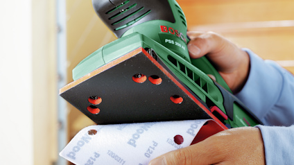 Person justerar sandpapper på en grön elektrisk slipmaskin märkt "Bosch".