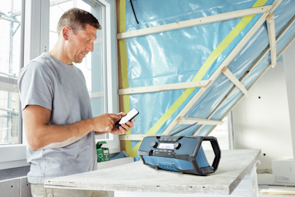 Man in a t-shirt using a smartphone next to a portable radio on a wooden workbench in a partially constructed room.