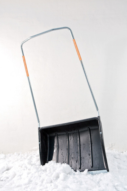 Snöskyffel med orange detaljer på handtaget, lutad mot en vit vägg, stående i snö.