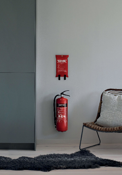 Brandsläckare och brandfilt monterade på en vit vägg bredvid en stol och grå matta.