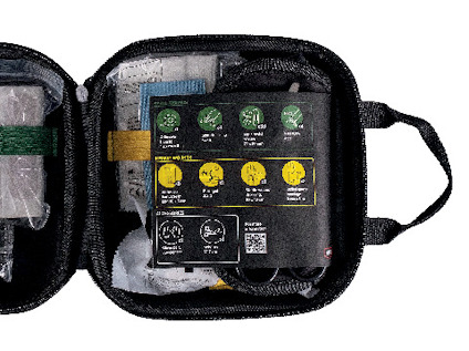 Öppen svart väska med medicinska förnödenheter, innehållande bandage, kompresser och instruktioner på locket.